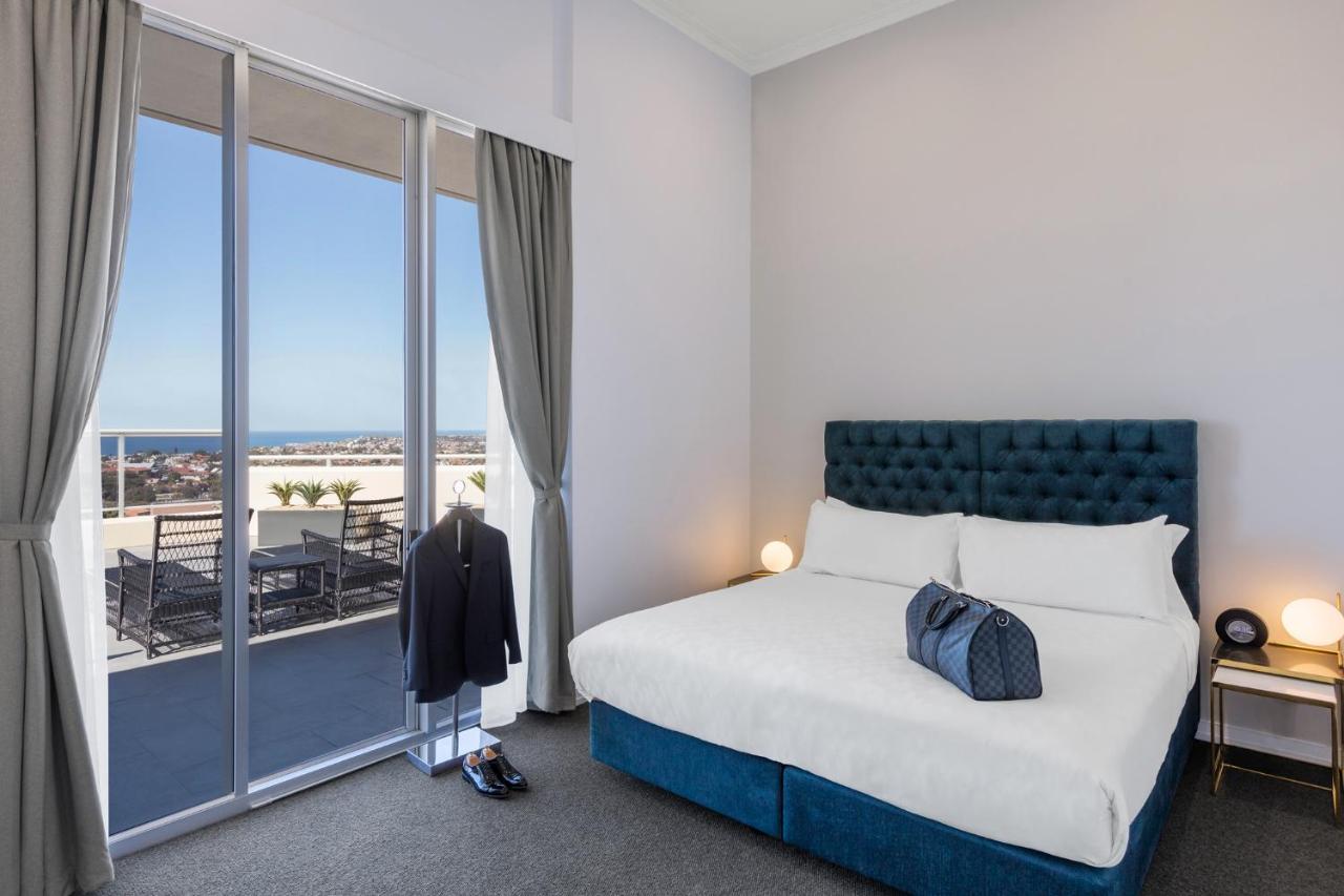 邦迪交界处美利通公寓式酒店 悉尼 外观 照片 The photo shows a stylish bedroom featuring a large bed with a tufted blue headboard. There's a neatly organized outfit consisting of a suit hanging on a stand, along with shoes placed on the floor. The room has large windows that provide a view of t