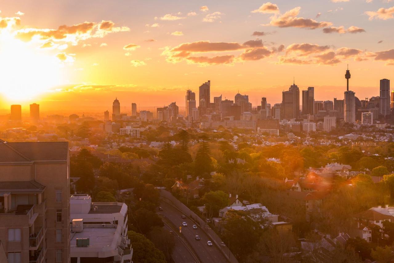 邦迪交界处美利通公寓式酒店 悉尼 外观 照片 The photo shows a panoramic view of a city skyline at sunset. The sun is low on the horizon, casting a warm golden light across the buildings. Tall skyscrapers are silhouetted against the colorful sky, which features hues of orange, pink, and yellow,