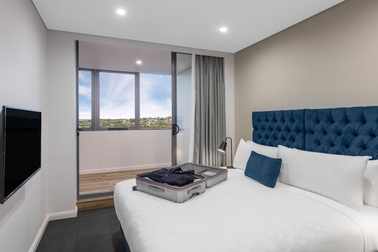 邦迪交界处美利通公寓式酒店 悉尼 外观 照片 The photo shows a modern hotel room featuring a double bed with a tufted blue headboard. There is a neatly arranged suitcase placed open on the bed, indicating that someone may be unpacking. The room has light-colored walls and a window allowing natu
