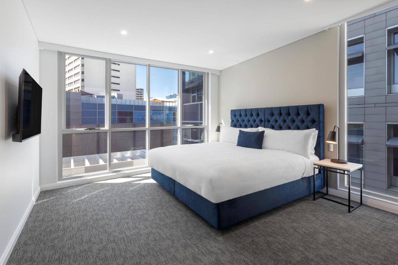 邦迪交界处美利通公寓式酒店 悉尼 外观 照片 The image shows a modern hotel room with a large, comfortable bed featuring a blue upholstered headboard. The room has large windows that allow natural light to enter, providing a good view of the surrounding buildings. There is a small bedside table