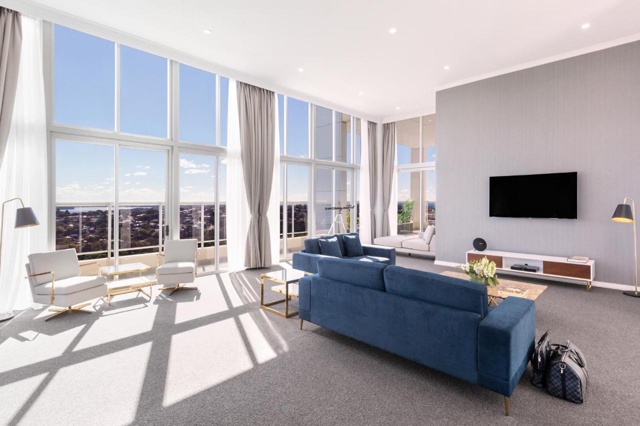 邦迪交界处美利通公寓式酒店 悉尼 外观 照片 The photo shows a bright and modern living room with large floor-to-ceiling windows that provide a view of the outdoors. The space features a blue sofa in the center, complemented by light-colored armchairs nearby. There are light curtains hanging by