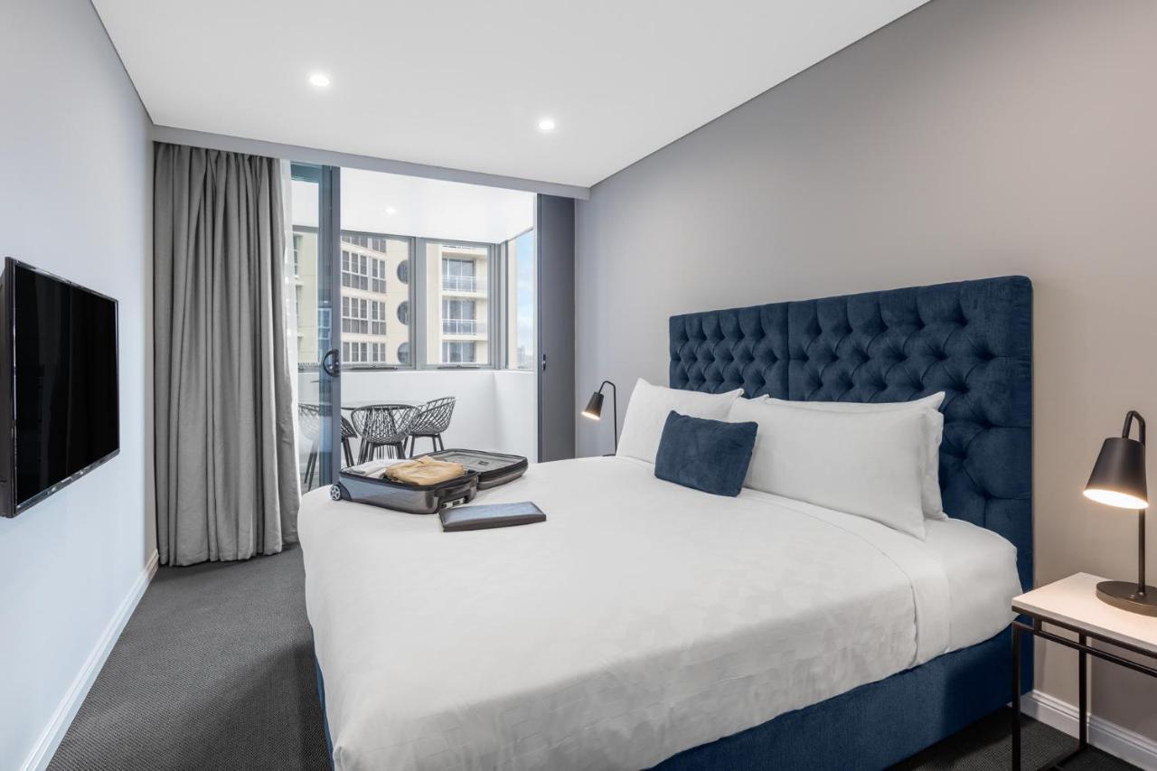 邦迪交界处美利通公寓式酒店 悉尼 外观 照片 The photo shows a modern hotel room featuring a large bed with a tufted blue headboard. There are minimalistic bedside tables with lamps on either side of the bed. A flat-screen TV is mounted on the wall across from the bed. To one side, there are fl