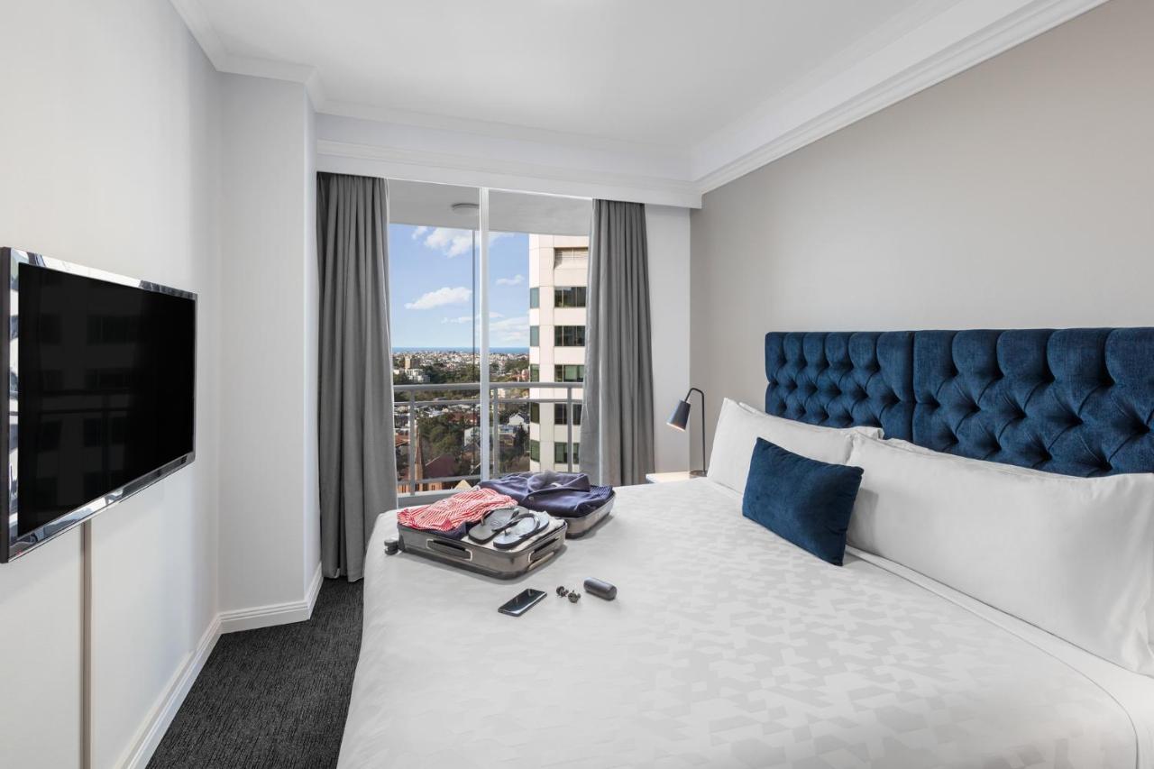 邦迪交界处美利通公寓式酒店 悉尼 外观 照片 The photo shows a modern hotel room featuring a neatly made bed with a blue tufted headboard and a white duvet. There's a small decorative pillow on the bed, and a piece of luggage is placed at the foot with clothes partially unpacked. A television i