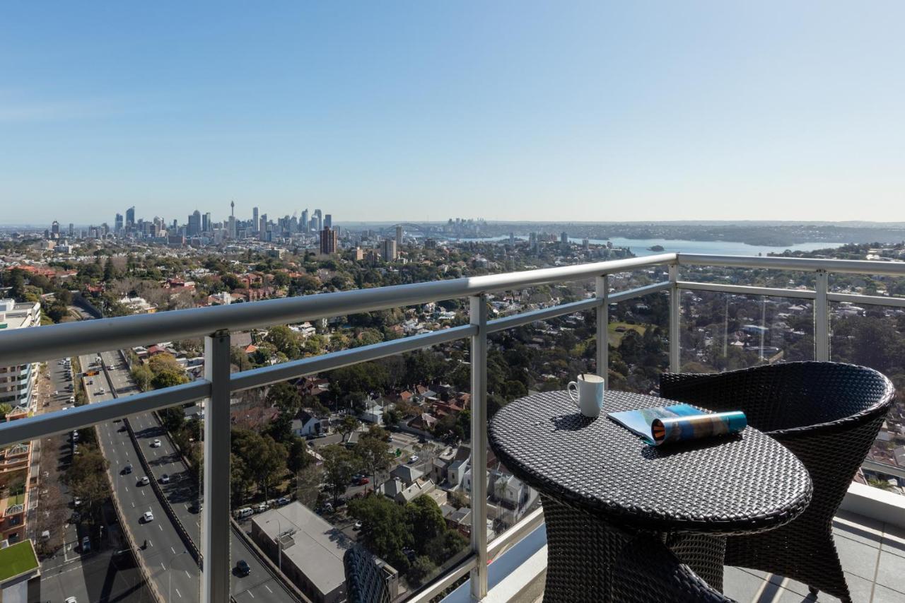 邦迪交界处美利通公寓式酒店 悉尼 外观 照片 The photo shows a balcony with a view overlooking a cityscape and a body of water in the distance. There is a round table with a woven design and two chairs, one of which has a mug on it. On the table, there’s also a rolled-up magazine or document. T