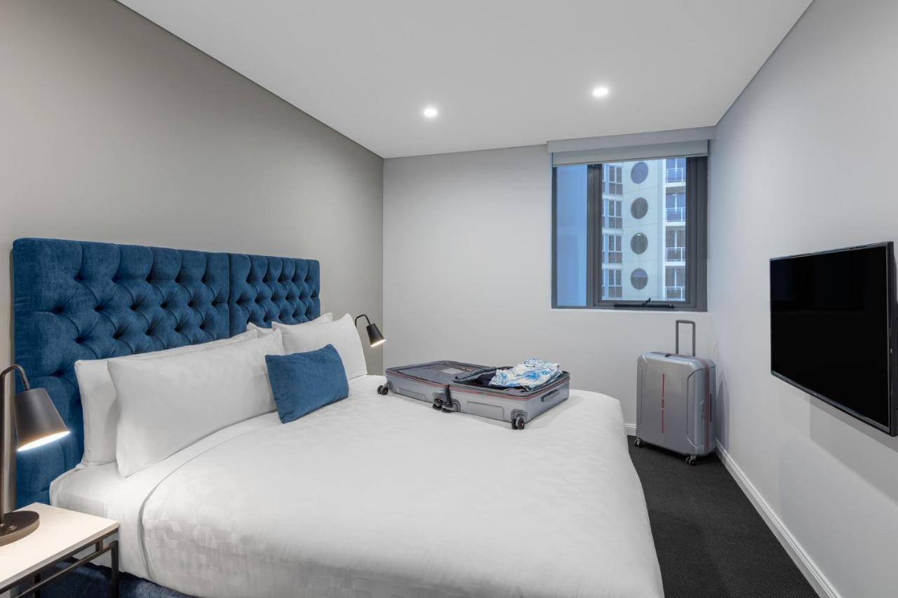 邦迪交界处美利通公寓式酒店 悉尼 外观 照片 The photo shows a modern hotel room featuring a neatly made bed with a blue tufted headboard. There is a small blue decorative pillow on the bed, and the bedding is white. Next to the bed, there is a nightstand with a lamp. A suitcase is partially op