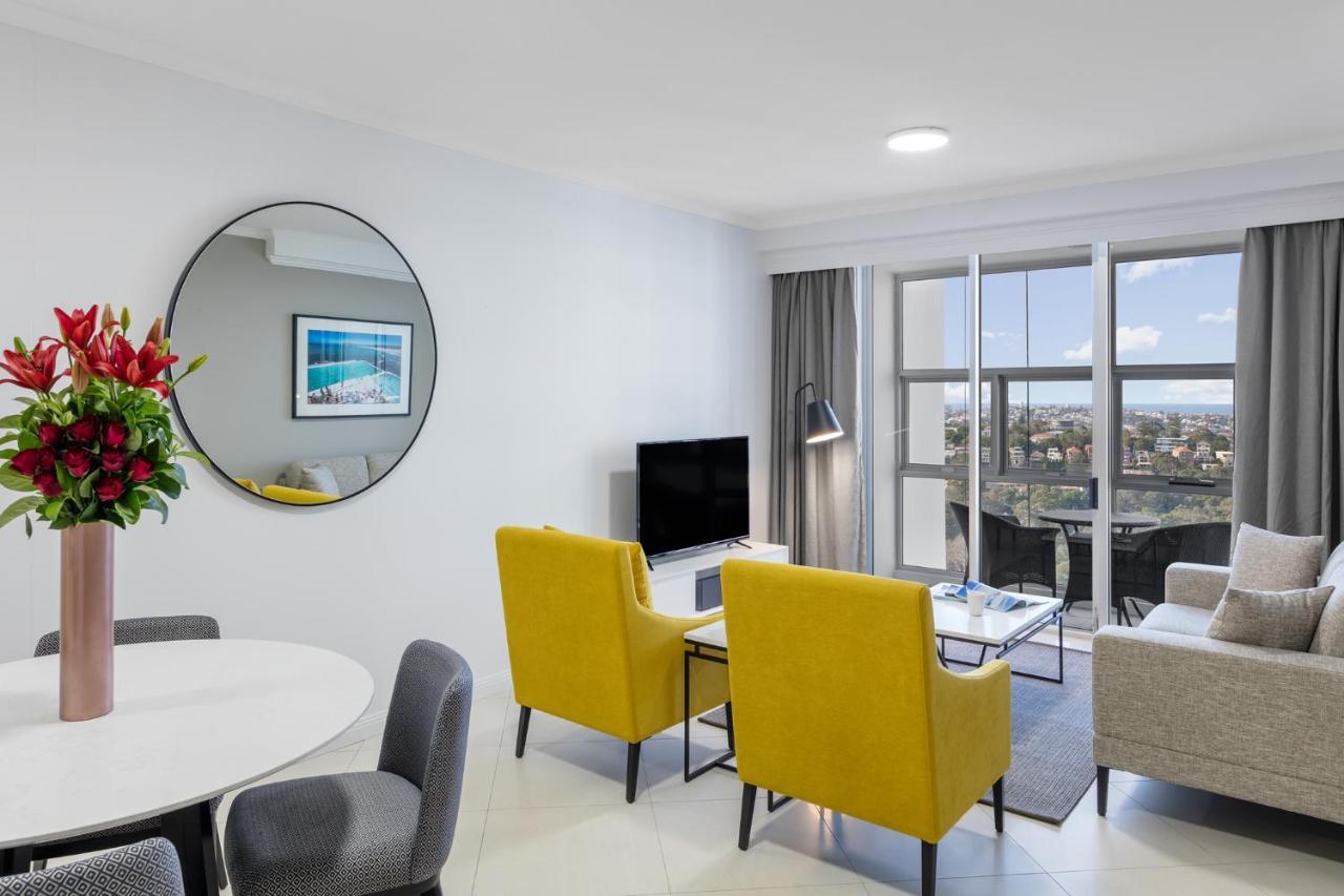 邦迪交界处美利通公寓式酒店 悉尼 外观 照片 The photo shows a modern living space with a stylish design. There are two bright yellow chairs facing a small glass table, and a comfortable beige sofa to the right. A TV is positioned on the wall in front of the chairs. To the left, there's a dinin