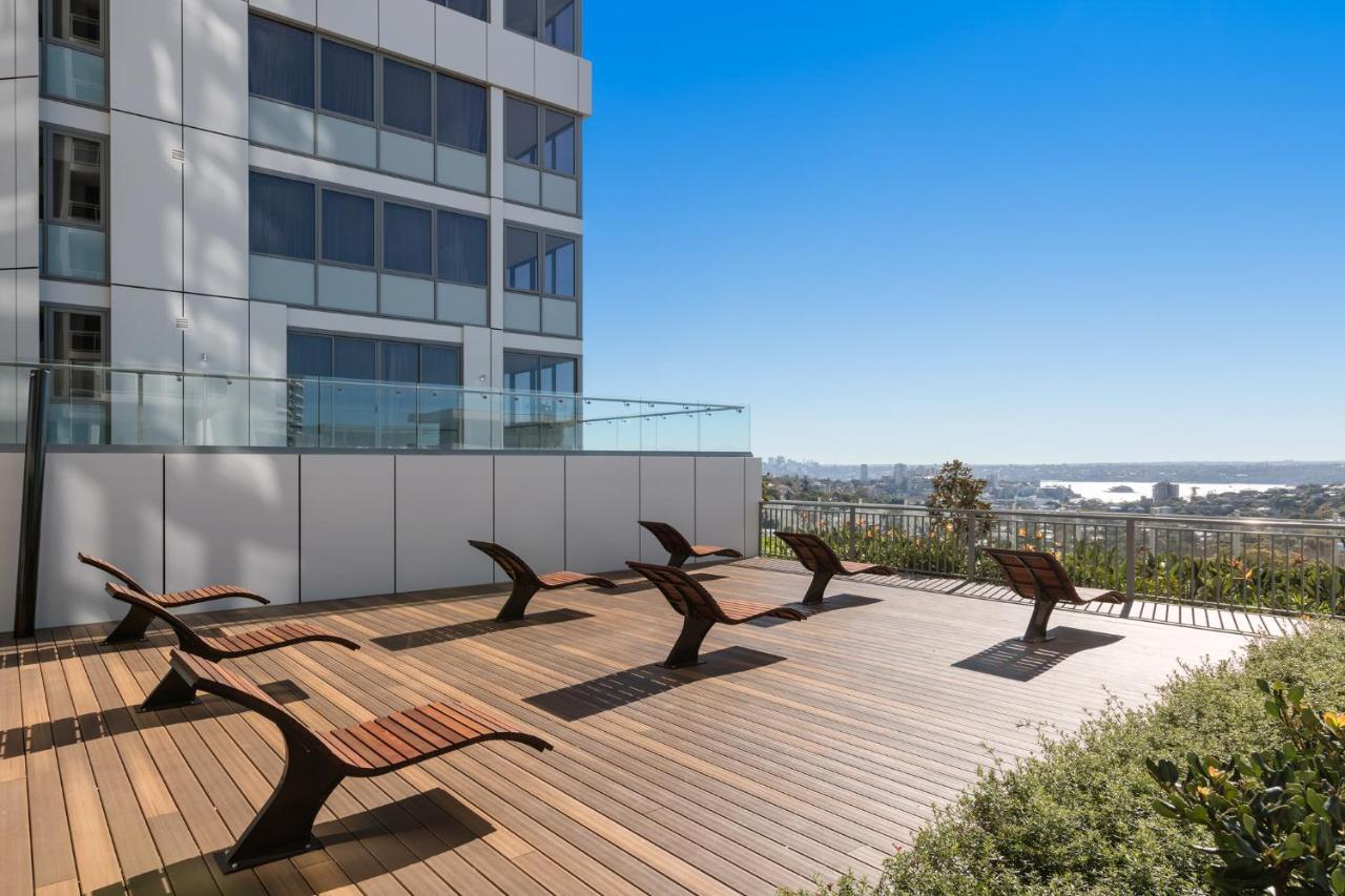 邦迪交界处美利通公寓式酒店 悉尼 外观 照片 The photo shows a rooftop or outdoor area with a wooden deck. There are several modern lounge chairs arranged neatly, facing a clear sky and a panoramic view of the surrounding landscape. In the background, there is a tall building with large windows