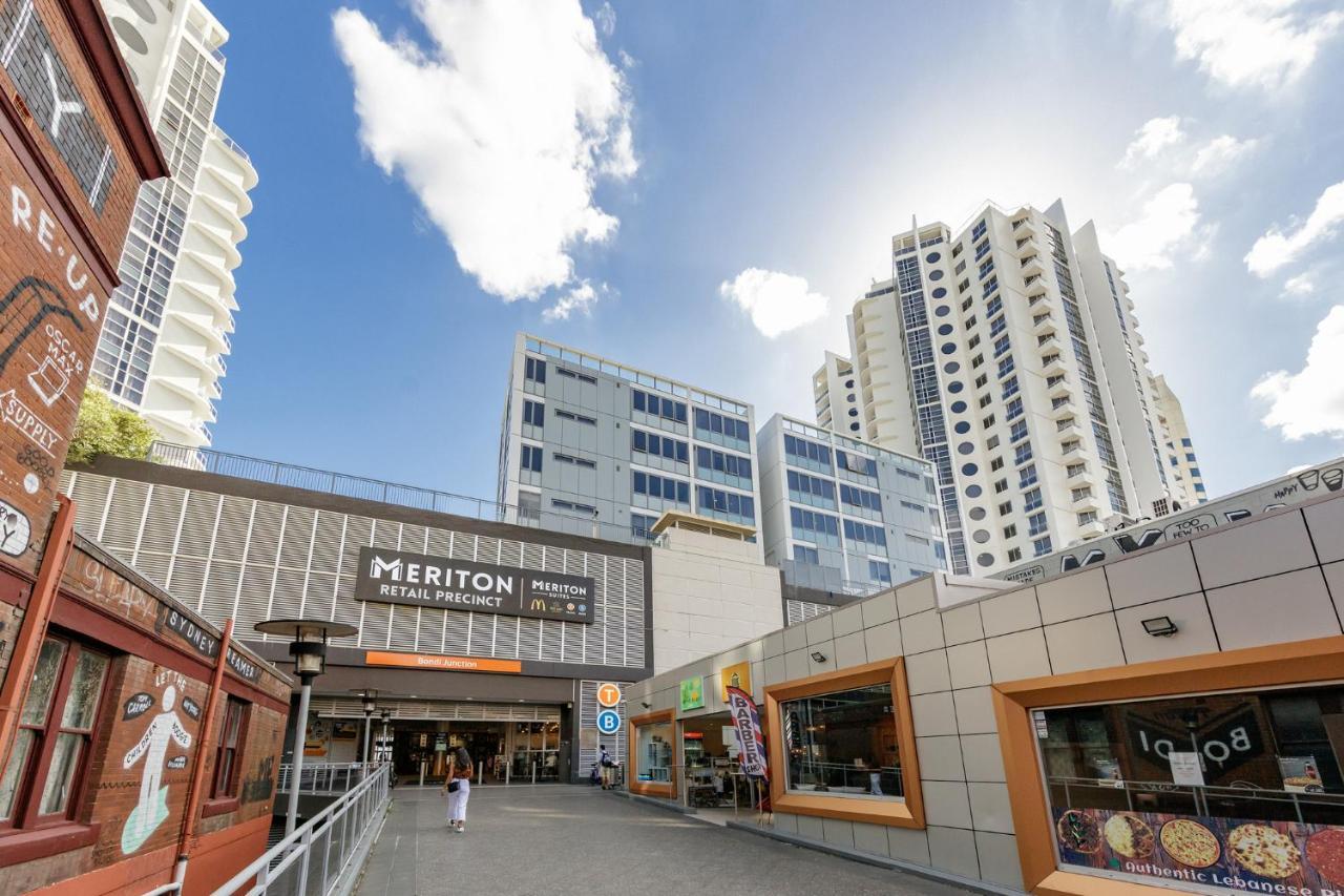 邦迪交界处美利通公寓式酒店 悉尼 外观 照片 The photo depicts a modern urban environment featuring a building labeled "Meriton Retail Precinct." In the foreground, there are shops with colorful signage, while in the background, tall residential buildings tower over the area. The sky above is b