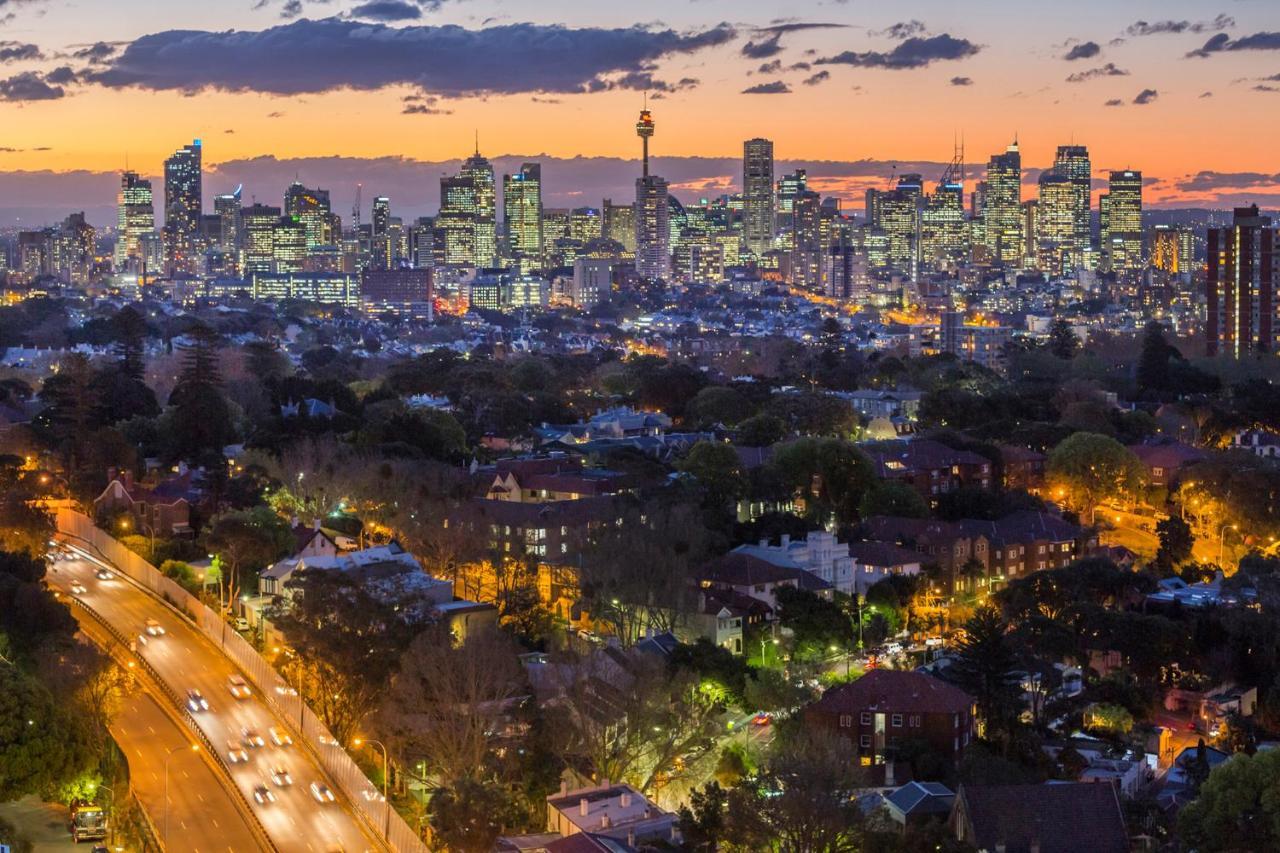 邦迪交界处美利通公寓式酒店 悉尼 外观 照片 The photo captures a stunning city skyline at dusk, featuring towering skyscrapers adorned with lights against a colorful sunset backdrop. In the foreground, a green area with trees and houses is visible, while a road with cars can be seen winding th