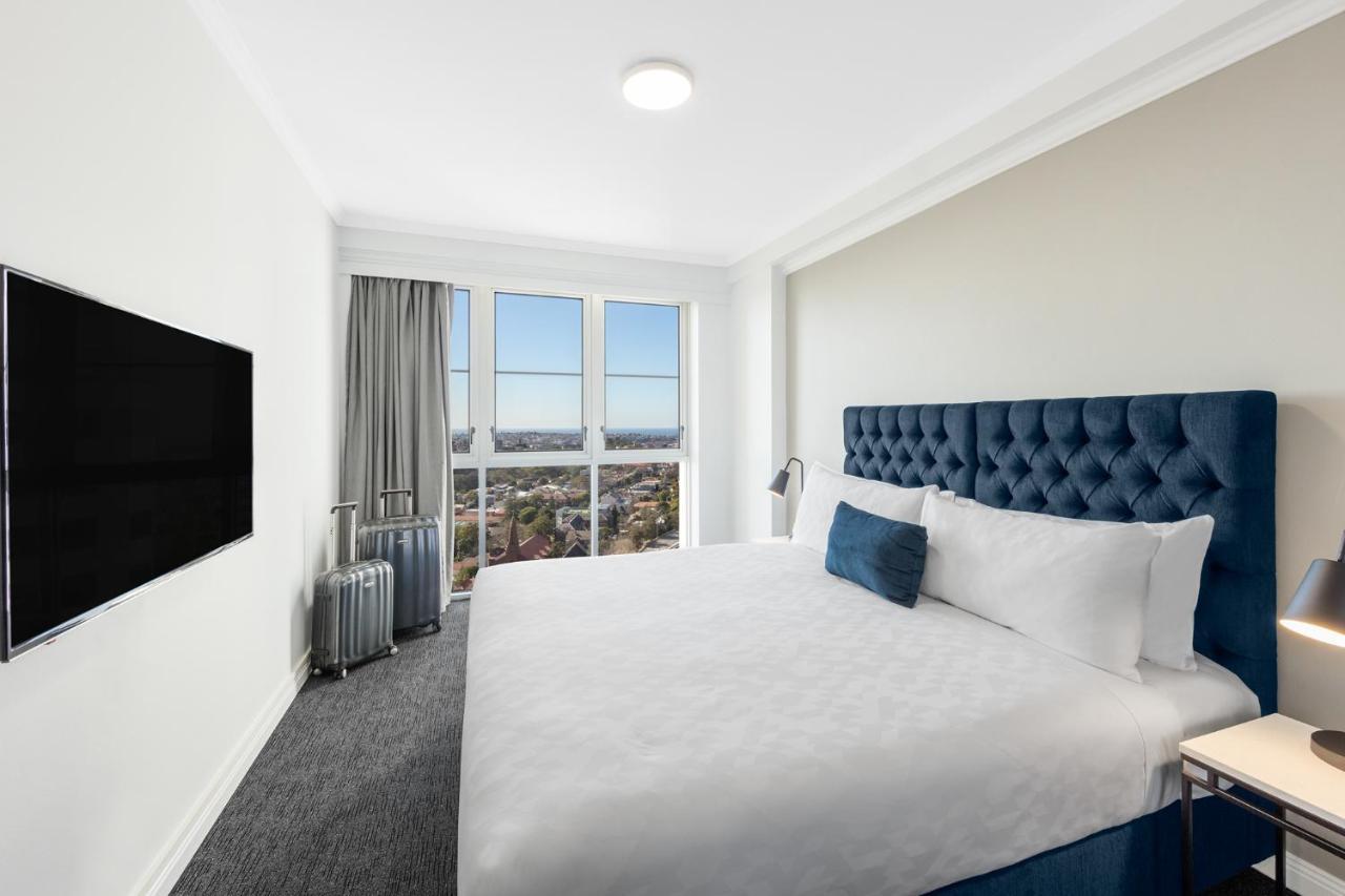 邦迪交界处美利通公寓式酒店 悉尼 外观 照片 The photo shows a modern hotel room with a large bed featuring a tufted blue headboard. There is a white duvet and a decorative pillow on the bed. A flat-screen television is mounted on the wall opposite the bed. Large windows are covered with gray c