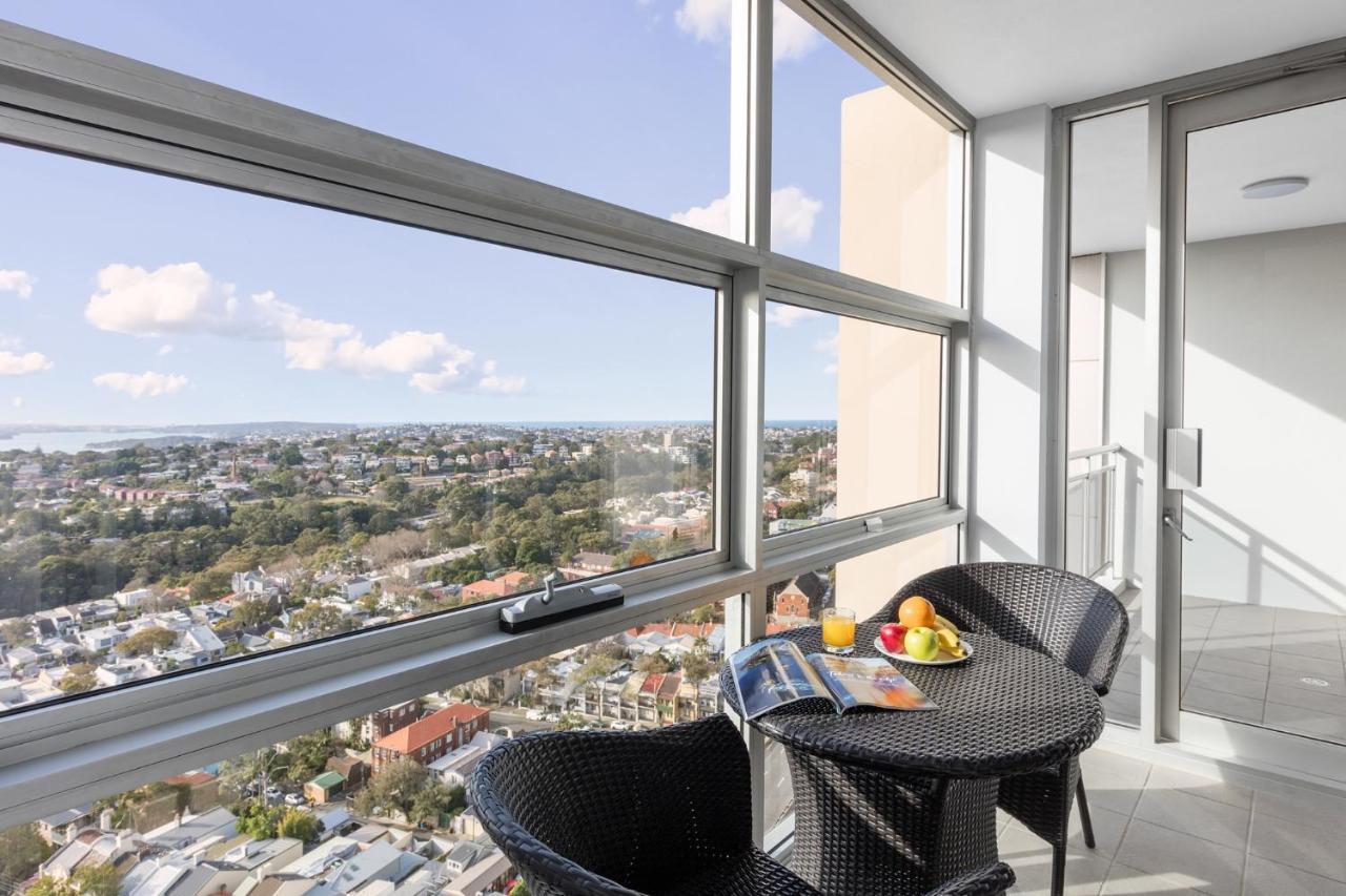 邦迪交界处美利通公寓式酒店 悉尼 外观 照片 The photo shows a modern balcony with large glass windows that offer a panoramic view of a sprawling urban landscape. There is a small round table made of dark wicker, accompanied by two matching chairs. On the table, there is a bowl of fruit and a m