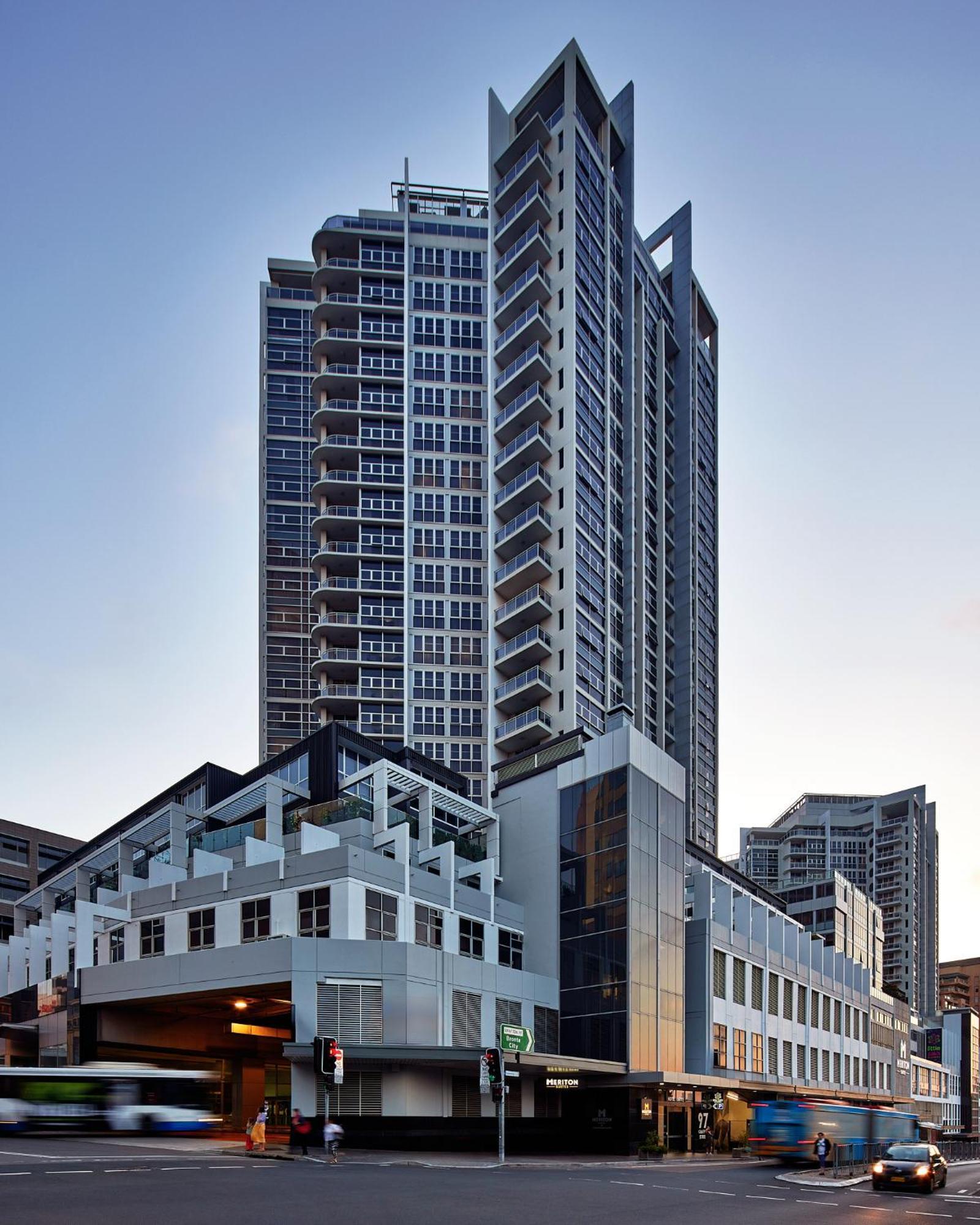 邦迪交界处美利通公寓式酒店 悉尼 外观 照片 The image shows a modern high-rise building with a sleek design. The structure features a mix of glass and concrete elements, with multiple levels showcasing balconies. The building appears to be part of an urban environment, likely containing reside
