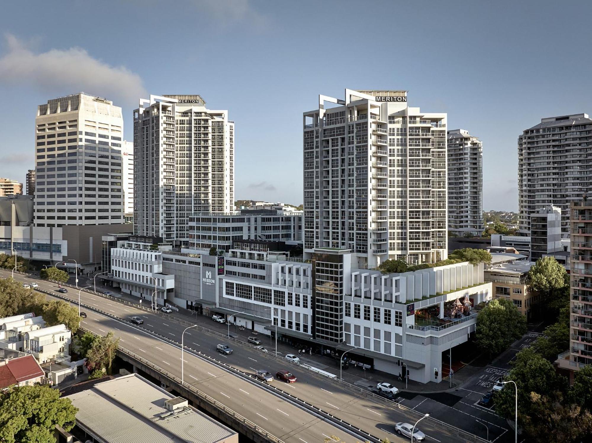 邦迪交界处美利通公寓式酒店 悉尼 外观 照片 The photo shows a cityscape featuring several tall modern buildings, likely apartments or office complexes. In the foreground, there is a road lined with cars, and the architecture of the buildings varies, with some having sleek glass facades and oth