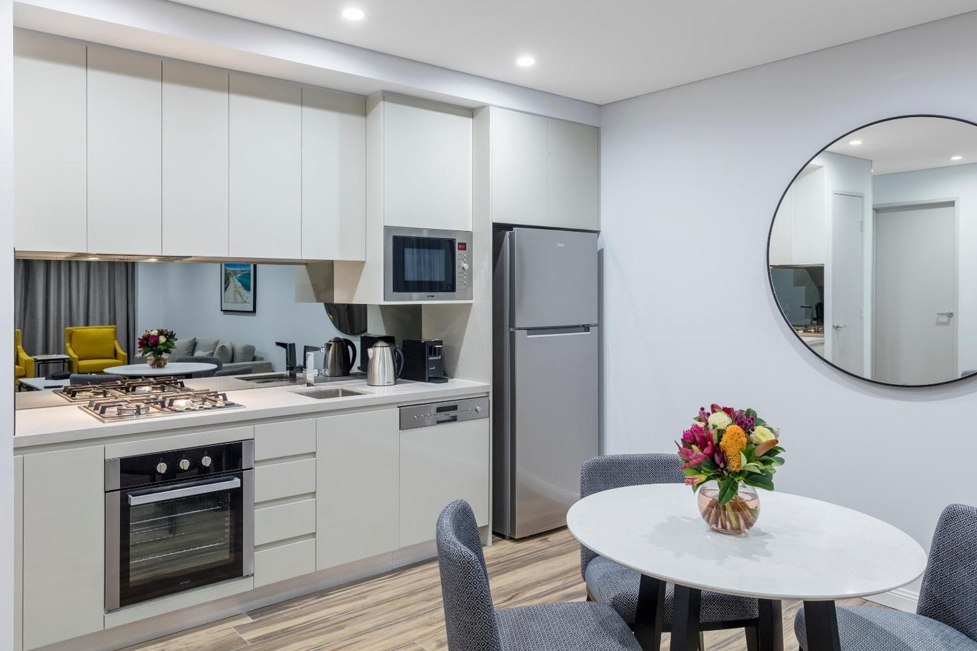 邦迪交界处美利通公寓式酒店 悉尼 外观 照片 The photo shows a modern kitchen and dining area. The kitchen features light-colored cabinetry with a built-in oven and stovetop, a microwave, and a silver refrigerator. There is a kettle and a coffee maker on the countertop. In the dining area, ther
